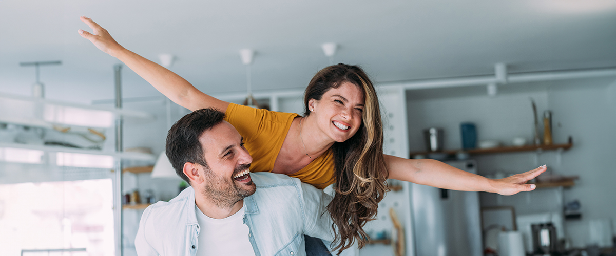 Das Bild zeigt ein fröhliches Paar in einer modernen, hellen Küche. Der Mann trägt ein helles Hemd und hebt die Frau auf seinem Rücken, während beide lachen. Die Frau hat langes, braunes Haar und trägt ein gelbes T-Shirt, sie streckt ihre Arme aus, als würde sie fliegen. Im Hintergrund sind Regale mit Küchengeräten und Dekorationen zu sehen. Das Bild vermittelt Freude und Leichtigkeit.