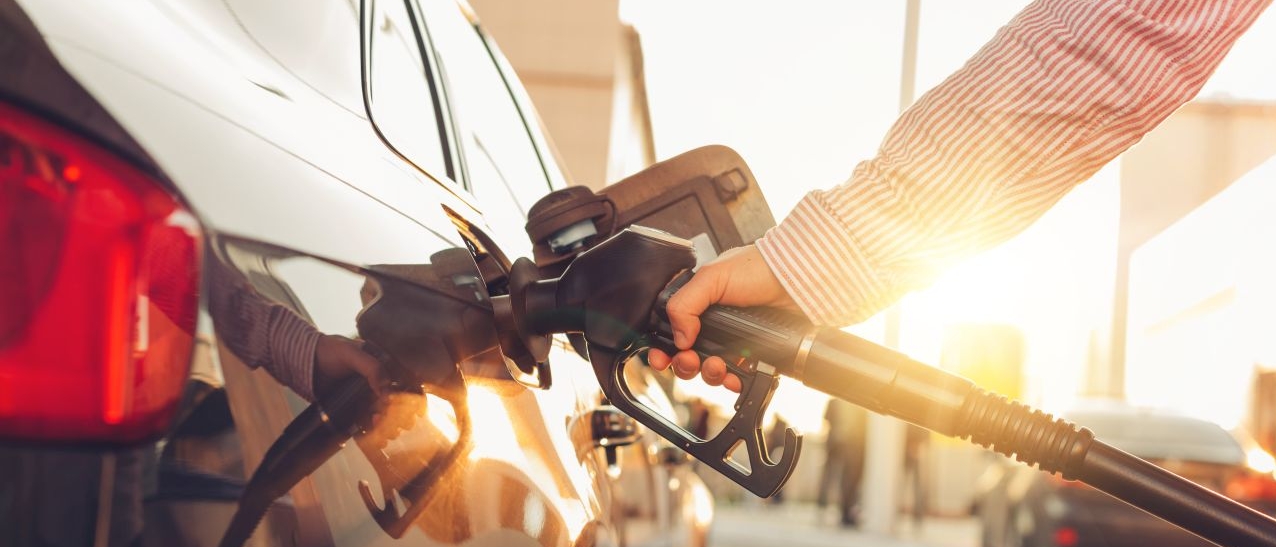Eine Person in einem gestreiften Hemd tankt ein Auto an einer Zapfsäule. Die Szene spielt sich bei Sonnenuntergang oder in der Abendsonne ab, was eine warme, goldene Lichtstimmung erzeugt. Der Fokus liegt auf der Hand, die den Zapfhahn hält, und dem Einfüllstutzen des Fahrzeugs. Im Hintergrund sind weitere Fahrzeuge und die typische Umgebung einer Tankstelle zu sehen.