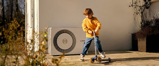 Ein Kind fährt mit einem Roller vor einem Haus, neben ihm ist die Außeneinheit einer Wärmepumpe installiert – modernes, energieeffizientes Heizsystem für nachhaltiges Heizen im Eigenheim.
