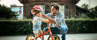 Ein Vater setzt seinem Kind einen Fahrradhelm auf, während es auf einem Laufrad sitzt – frühe Vorsorge und finanzielle Planung für Kinder als Basis für den langfristigen Vermögensaufbau.