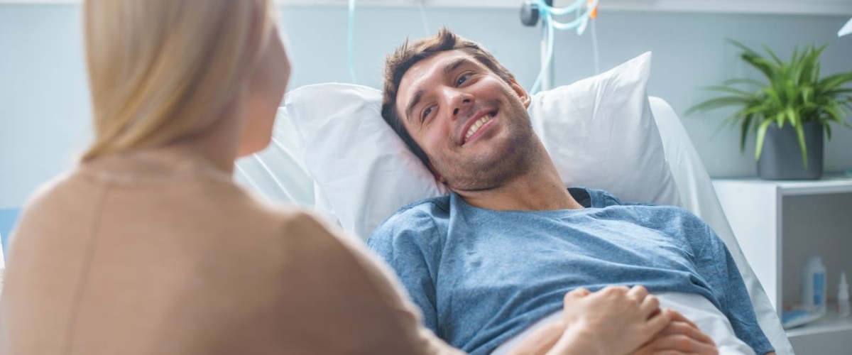 In the Hospital Sick Man Lying on the Bed, His Visiting Wife is Sitting Beside Him and Talking, Holding His Hands.