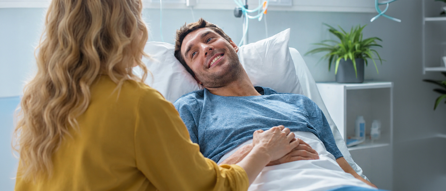 Ein Mann liegt nach einer Verletzung im Krankenhausbett, während eine Frau ihm unterstützend die Hand hält – Symbolbild für eine unerwartete Erkrankung oder einen Unfall vor der Reise und den Schutz durch eine Reiserücktrittsversicherung.