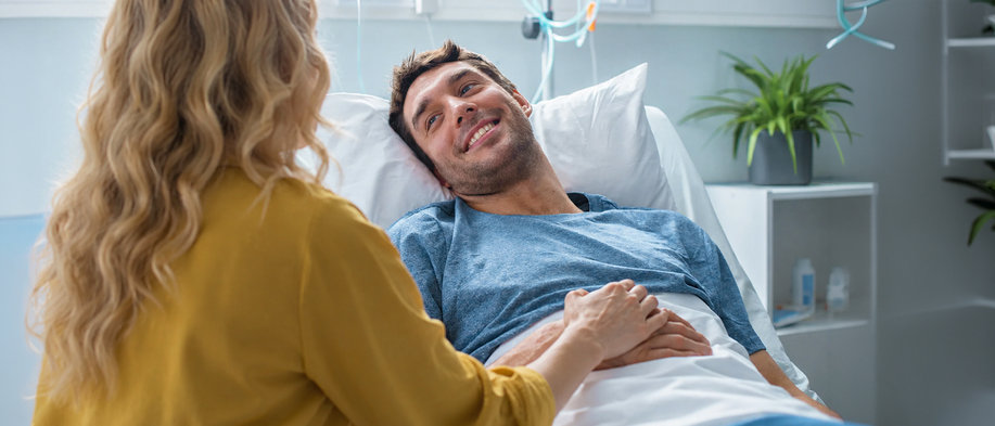 Ein Mann liegt nach einer Verletzung im Krankenhausbett, während eine Frau ihm unterstützend die Hand hält – Symbolbild für eine unerwartete Erkrankung oder einen Unfall vor der Reise und den Schutz durch eine Reiserücktrittsversicherung.