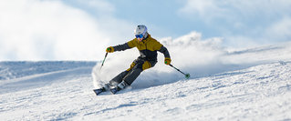 Skifahrer in goldener Kleidung fährt dynamisch eine verschneite Piste hinunter und wirbelt dabei Schnee auf, vor hellem Himmel und Berglandschaft.