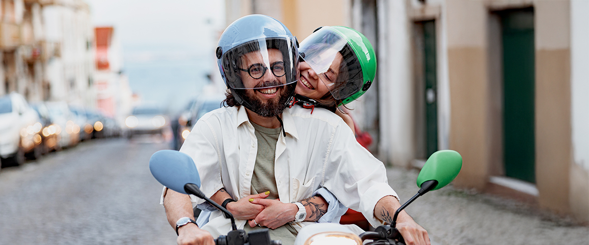 Zwei Erwachsene mit Schutzhelmen fahren gemeinsam auf einem Motorroller durch eine städtische Straße, sie lächeln und zeigen moderne, sichere urbane Mobilität.