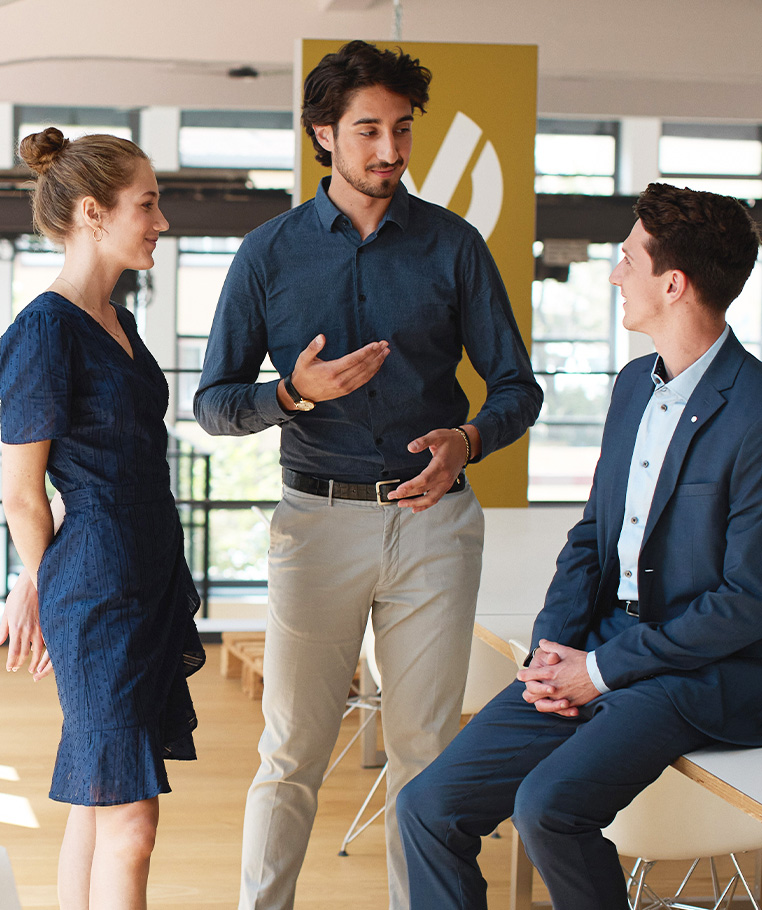 drei Finanzcoaches im Gespräch, eine blonde Frau und zwei dunkelhaarige Männer in legeren Businessoutfits