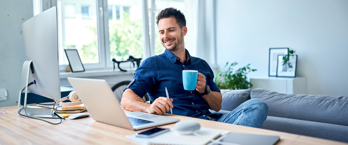 Mann am Schreibtisch, Kaffee und Kugelschreiber in der HAnd, auf dem Tisch Bildschirm und Laptop